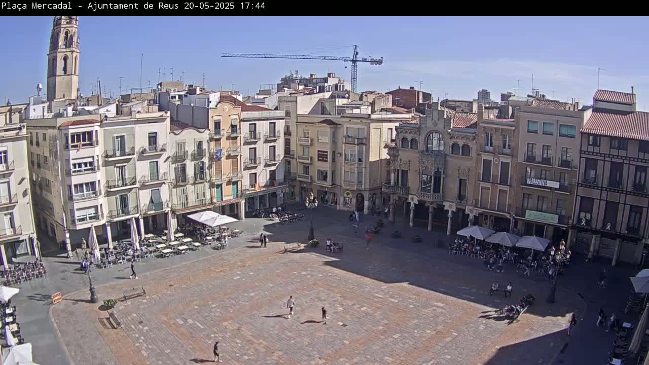 A large, paved town square is surrounded by multi-story buildings under a clear, sunny sky, with several people scattered throughout and some seated at outdoor cafes.