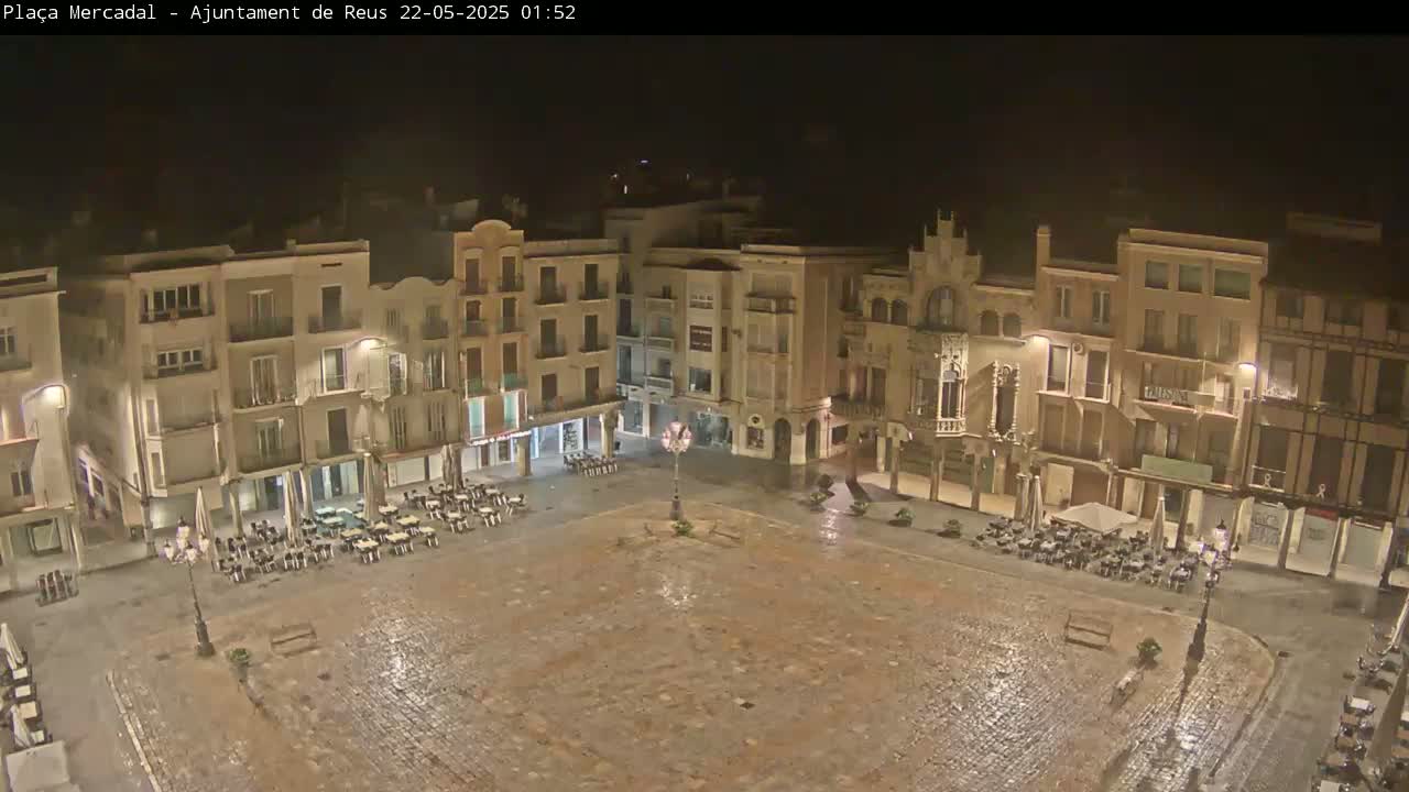 A wet, empty town square at night is surrounded by multi-story buildings with illuminated windows and sparsely occupied outdoor seating areas.