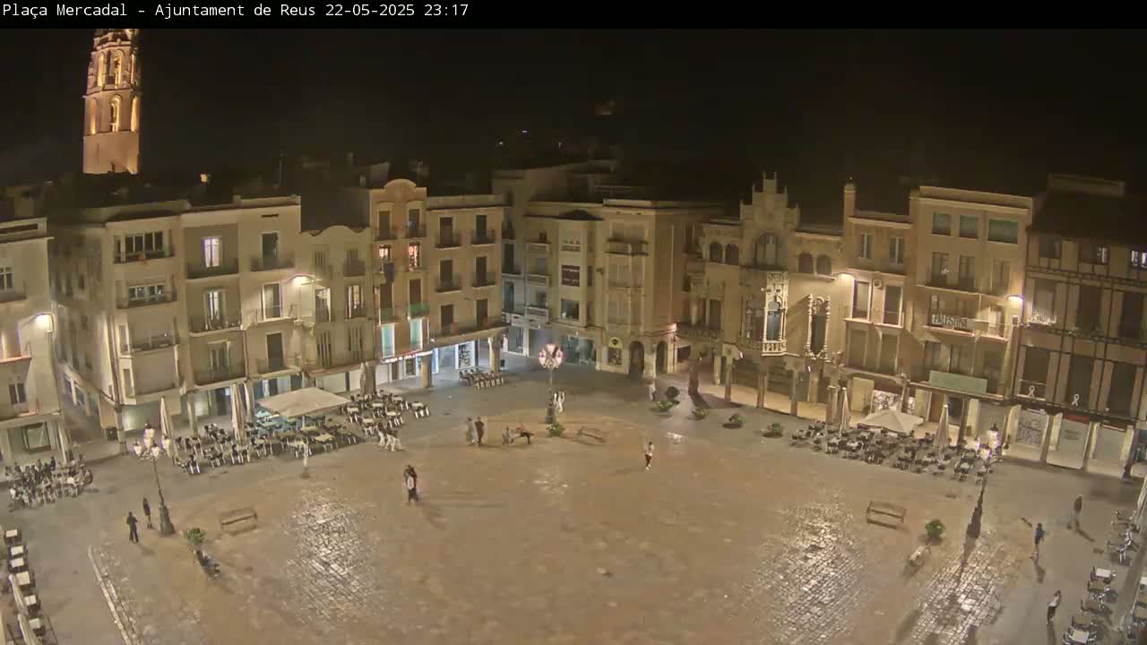 A nighttime, wet, cobblestone town square is sparsely populated with people and outdoor seating areas in front of illuminated buildings, one of which is a tall tower.