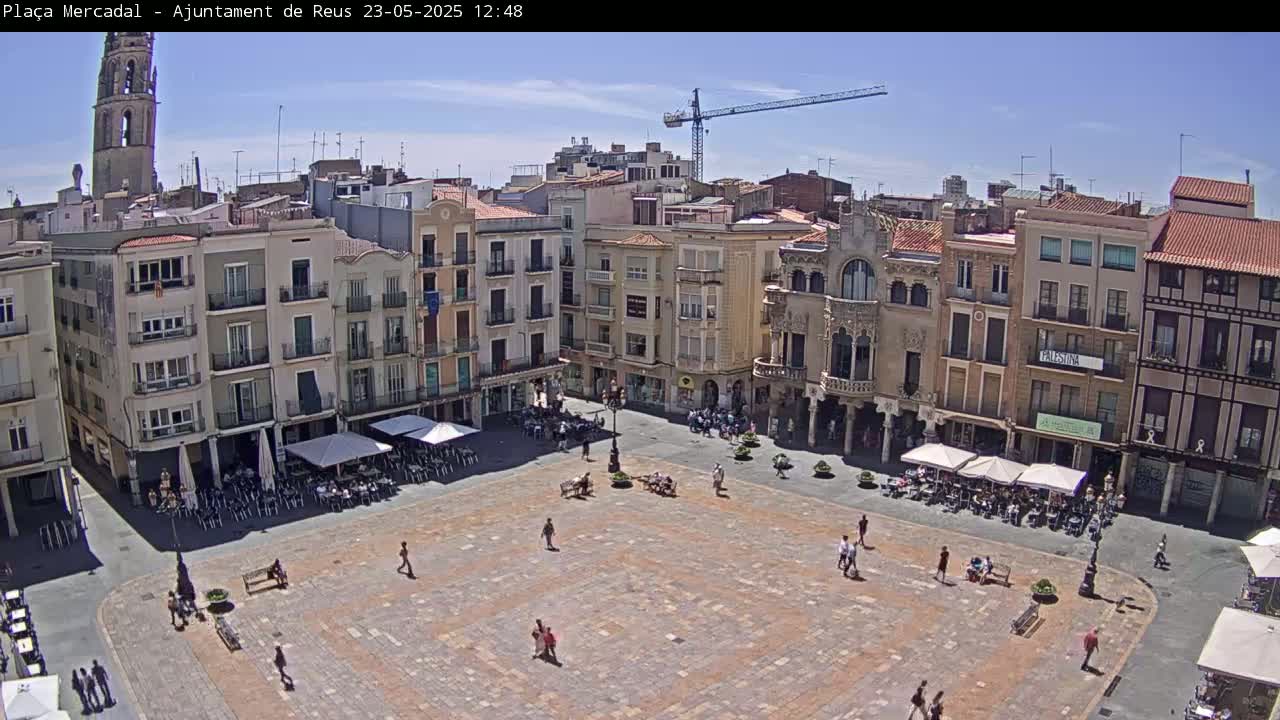A large, paved town square with several people and outdoor seating areas is surrounded by multi-story buildings under a clear, sunny sky.