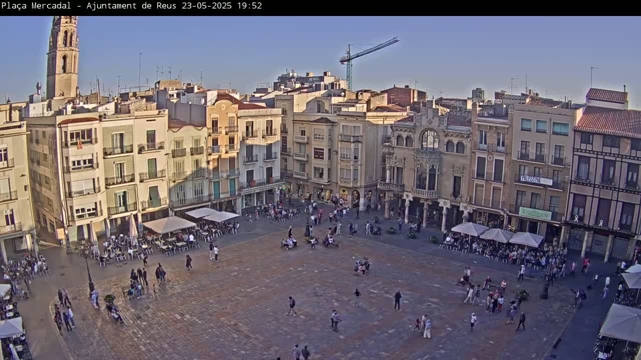 A large town square, filled with people and outdoor seating at cafes, is surrounded by multi-story buildings under a clear, late afternoon sky.
