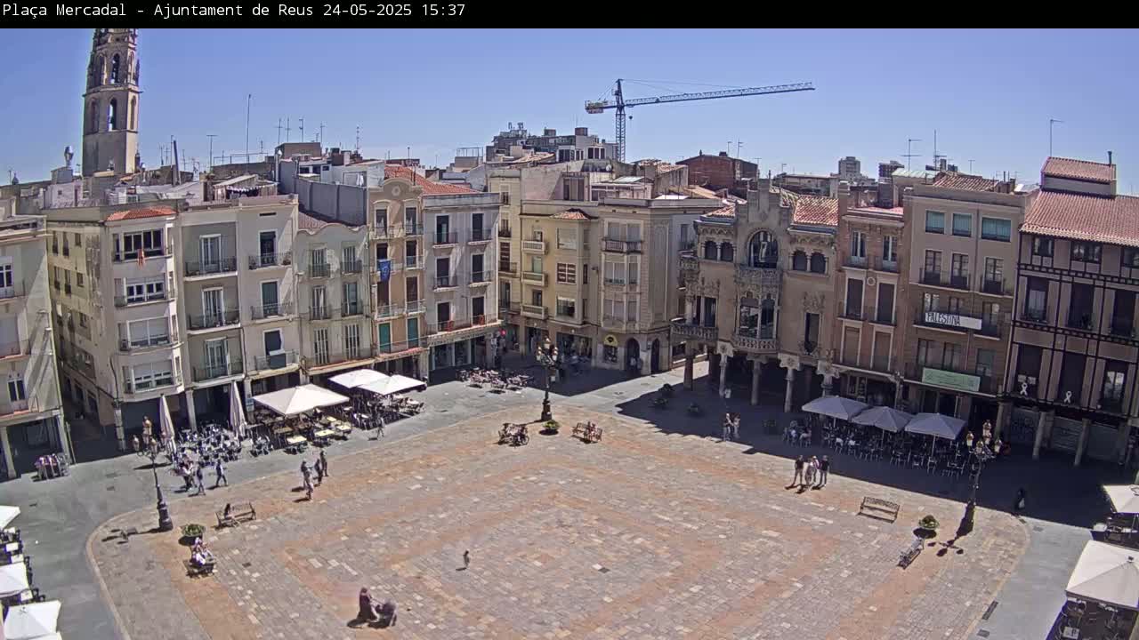 A large, paved town square with several buildings surrounding it, outdoor cafes with umbrellas, and a construction crane in the background under a clear, sunny sky.