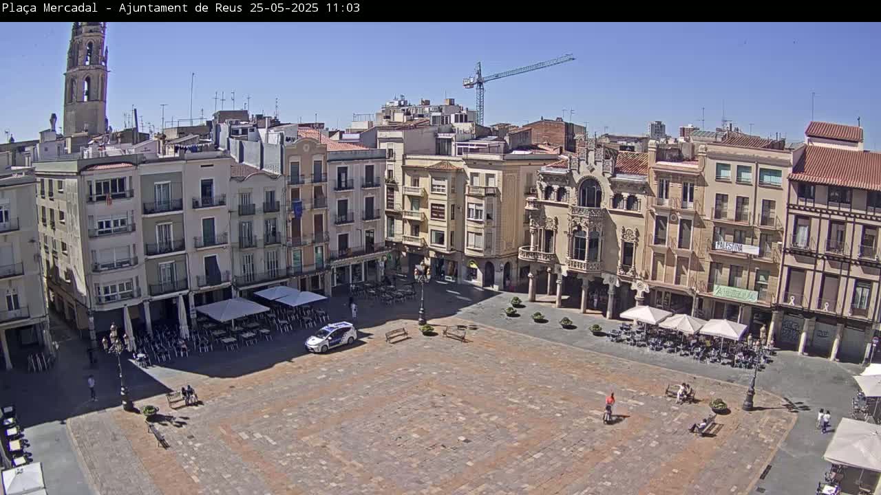 A large, mostly empty town square with surrounding buildings, outdoor cafes, and a police car under a clear, sunny sky.