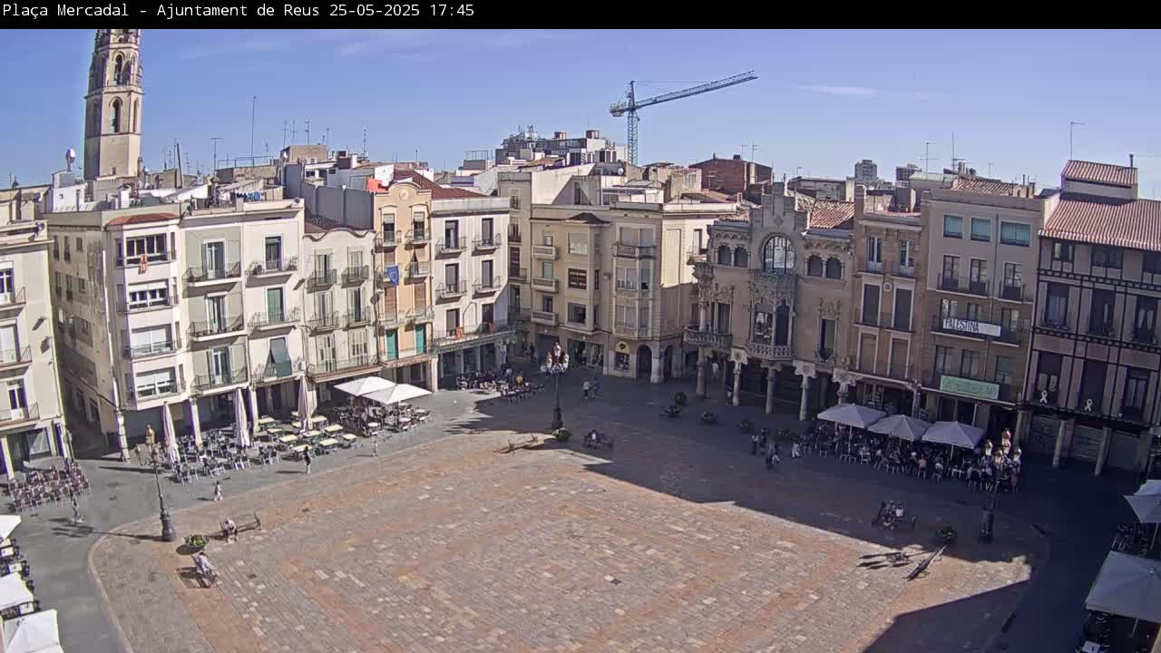 A large, mostly empty town square with several buildings surrounding it, outdoor seating areas with people, and a construction crane in the background under a clear, sunny sky.