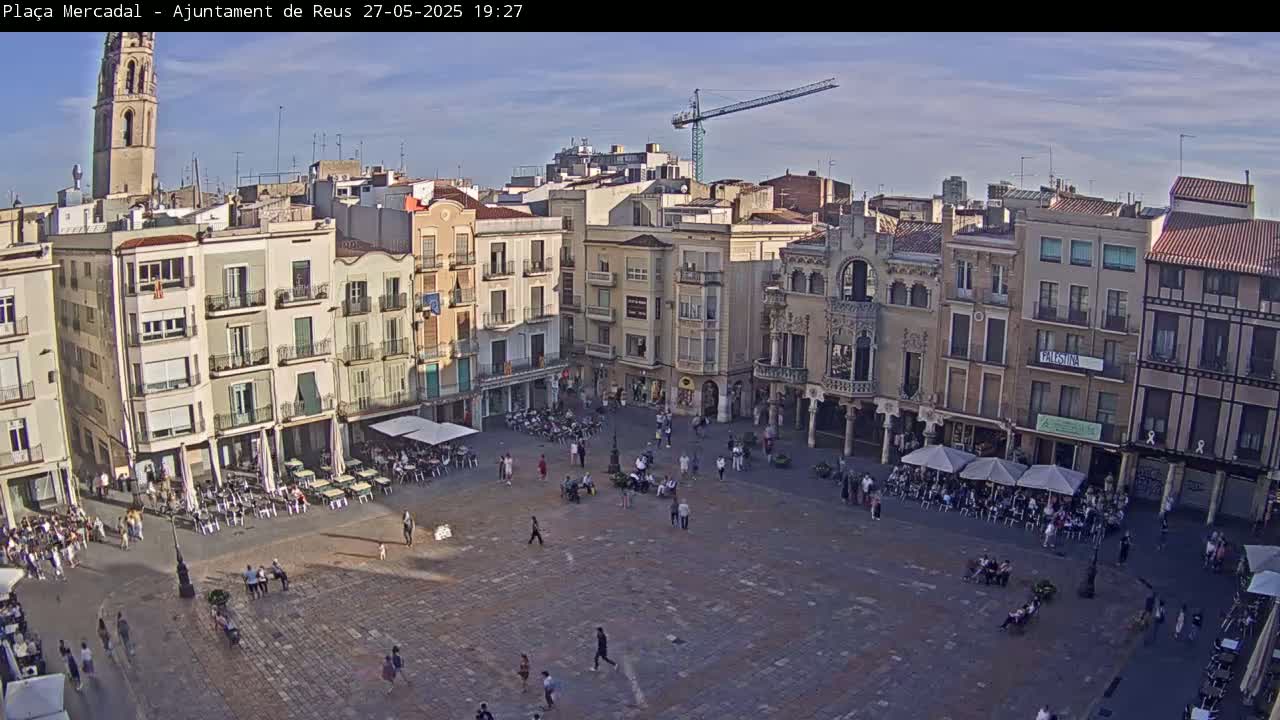 A large, mostly empty town square is surrounded by multi-story buildings under a mostly clear, sunny sky.