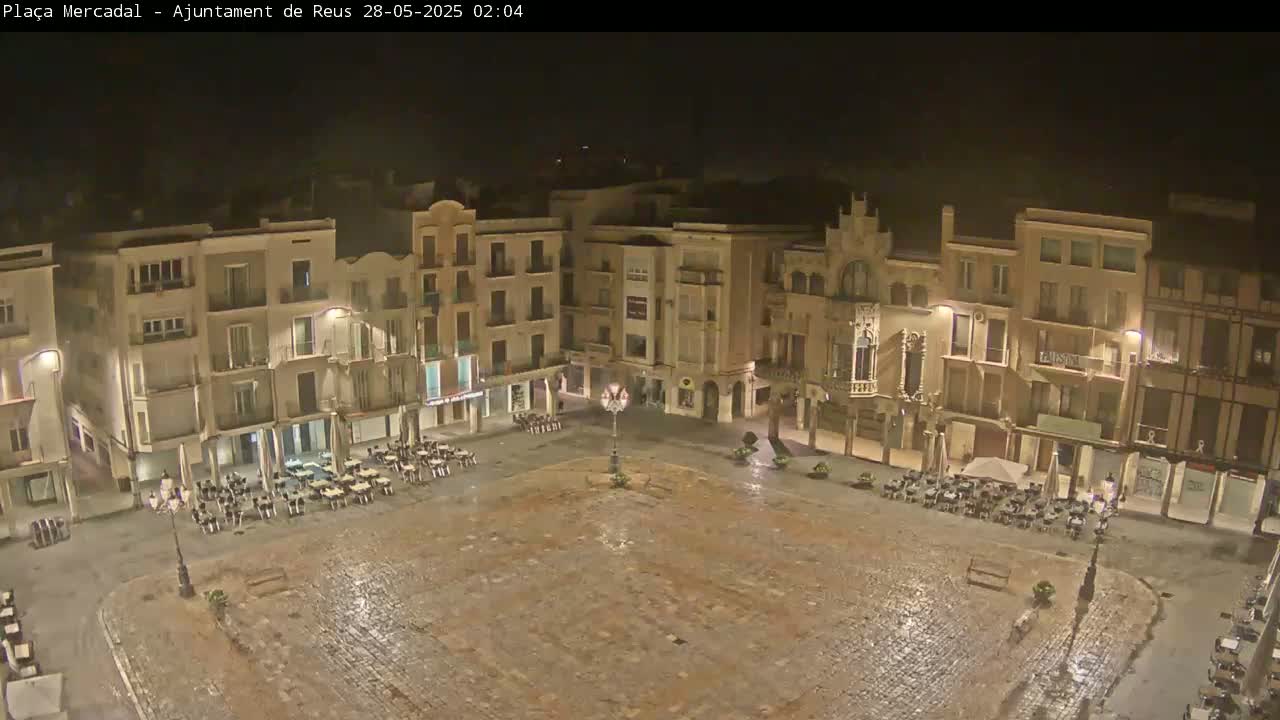 A nighttime, wet cobblestone town square is surrounded by multi-story buildings with sparsely occupied outdoor seating areas.