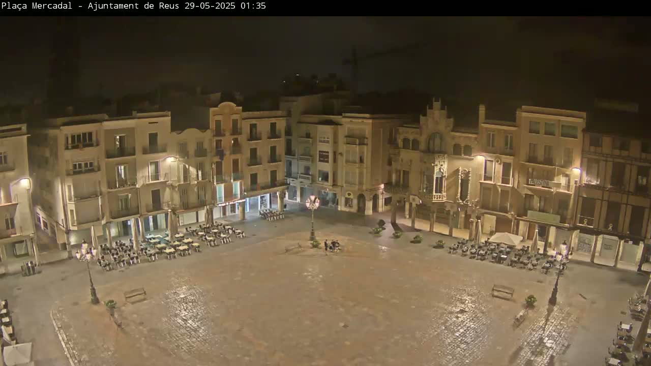 A wet, empty town square at night, surrounded by multi-story buildings with illuminated windows and sparsely occupied outdoor seating areas.
