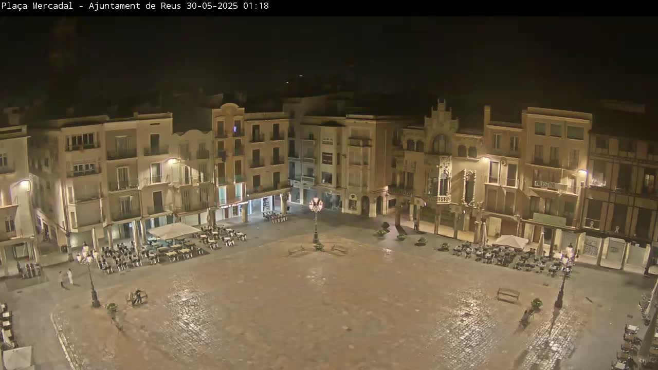 A wet, empty town square at night, surrounded by multi-story buildings with outdoor seating areas.