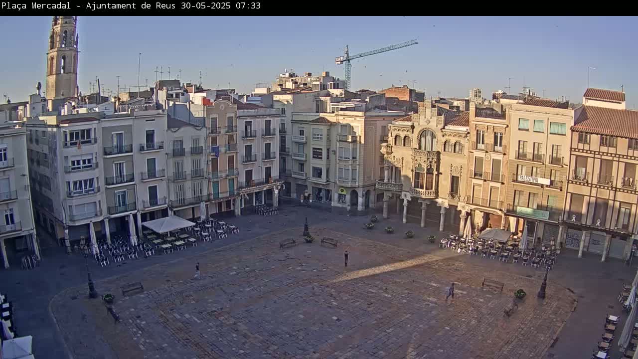 A large, mostly empty town square with surrounding buildings under a clear, bright sky, and a construction crane visible in the distance.