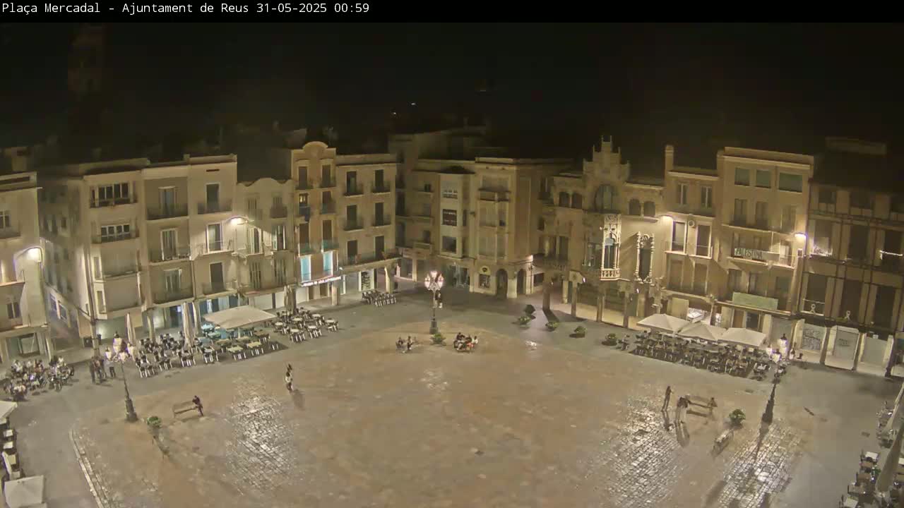 A mostly empty, cobblestone town square at night, surrounded by multi-story buildings with outdoor seating areas, is slightly wet from a recent rain.