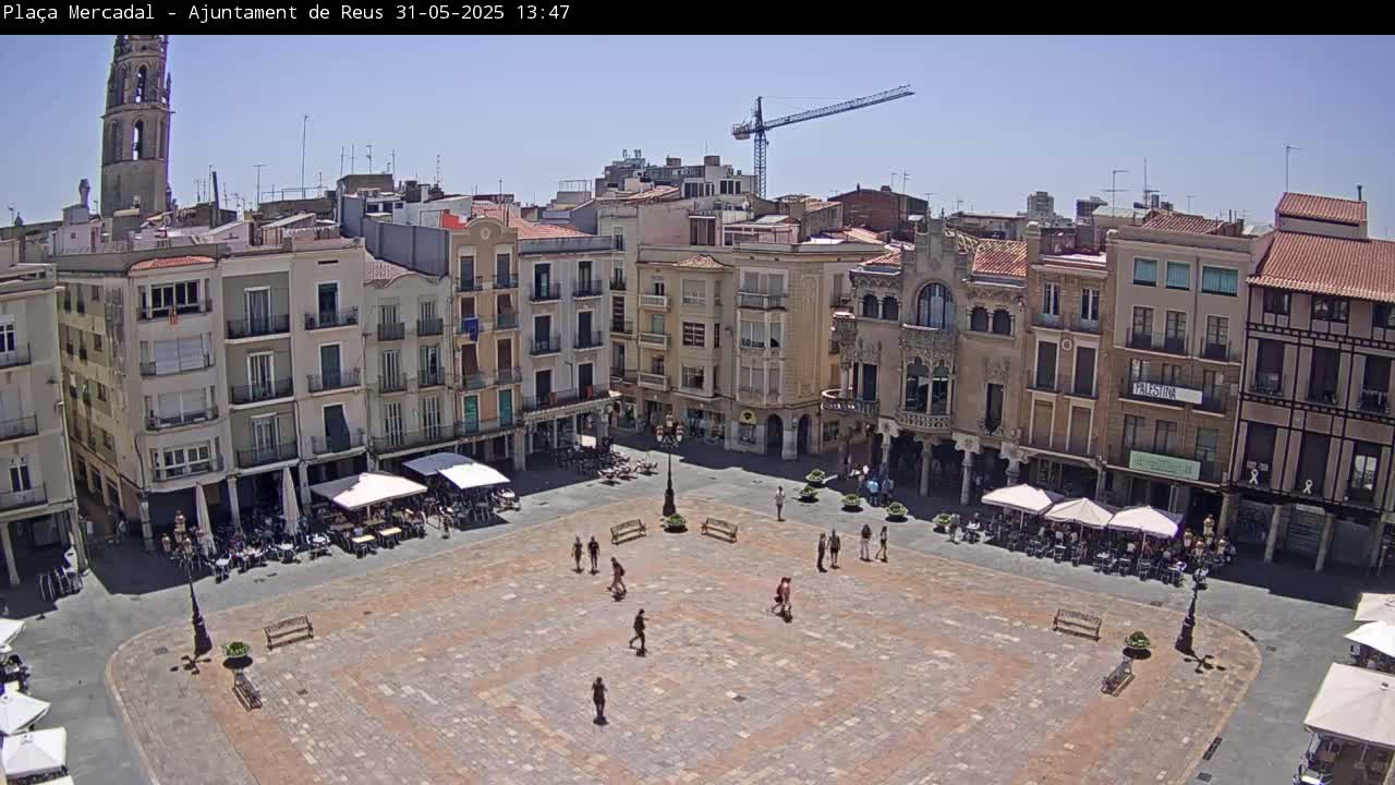 A large town square, under a clear sunny sky, is surrounded by multi-story buildings with outdoor cafes and a few pedestrians.