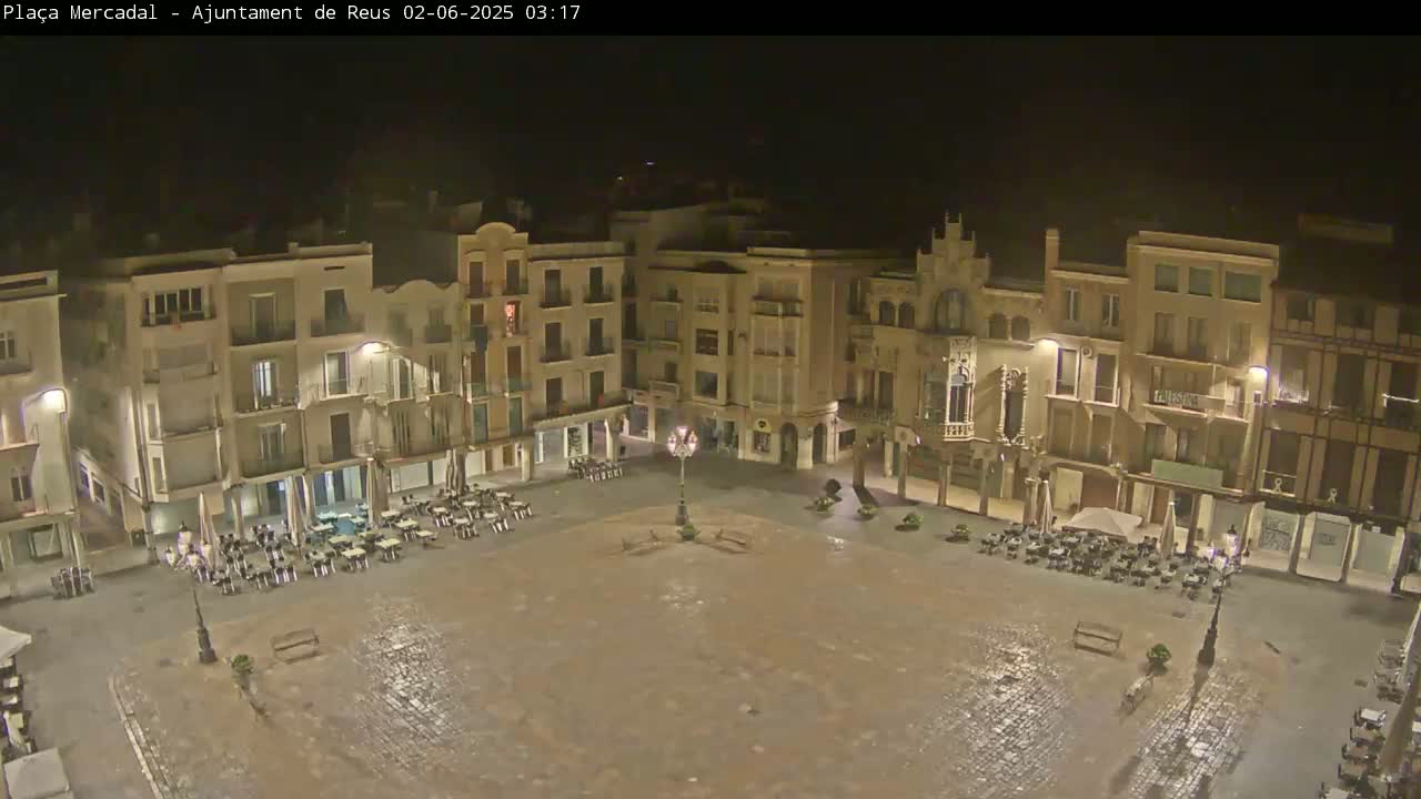 A nighttime, wet cobblestone plaza is surrounded by multi-story buildings with sparsely occupied outdoor seating areas.