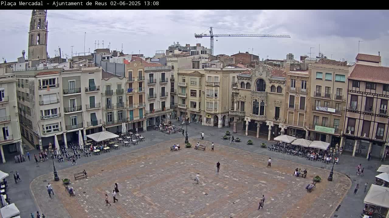 A large, paved town square with several buildings surrounding it, outdoor cafes with patrons, and a few people walking around, under an overcast sky.
