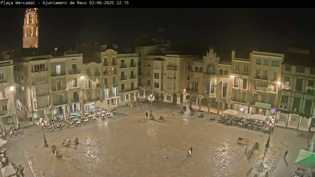 A nighttime, wet town square is surrounded by multi-story buildings, with some people sitting at outdoor tables and others walking around.