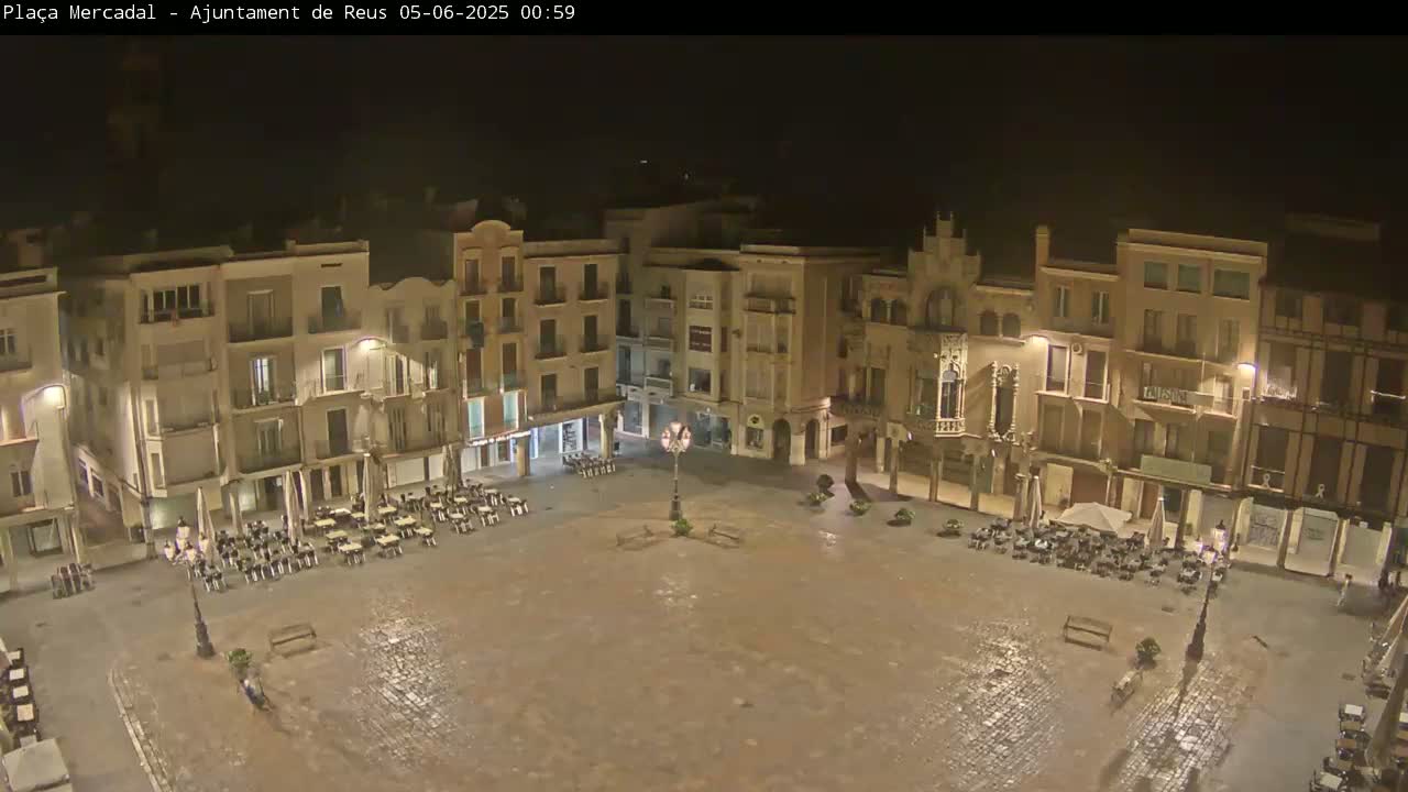 A nighttime, wet cobblestone town square is surrounded by multi-story buildings with outdoor seating areas.