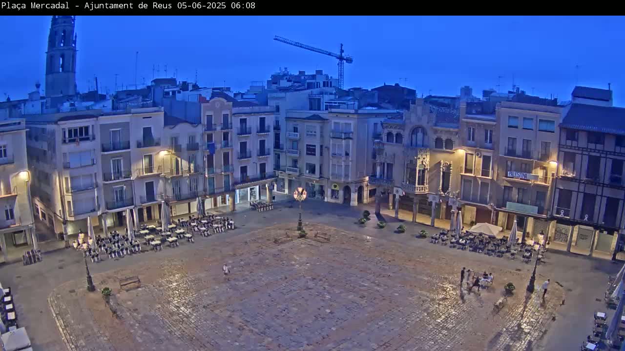 A large, mostly empty town square at twilight, surrounded by multi-story buildings, with a few people and outdoor cafe seating visible.