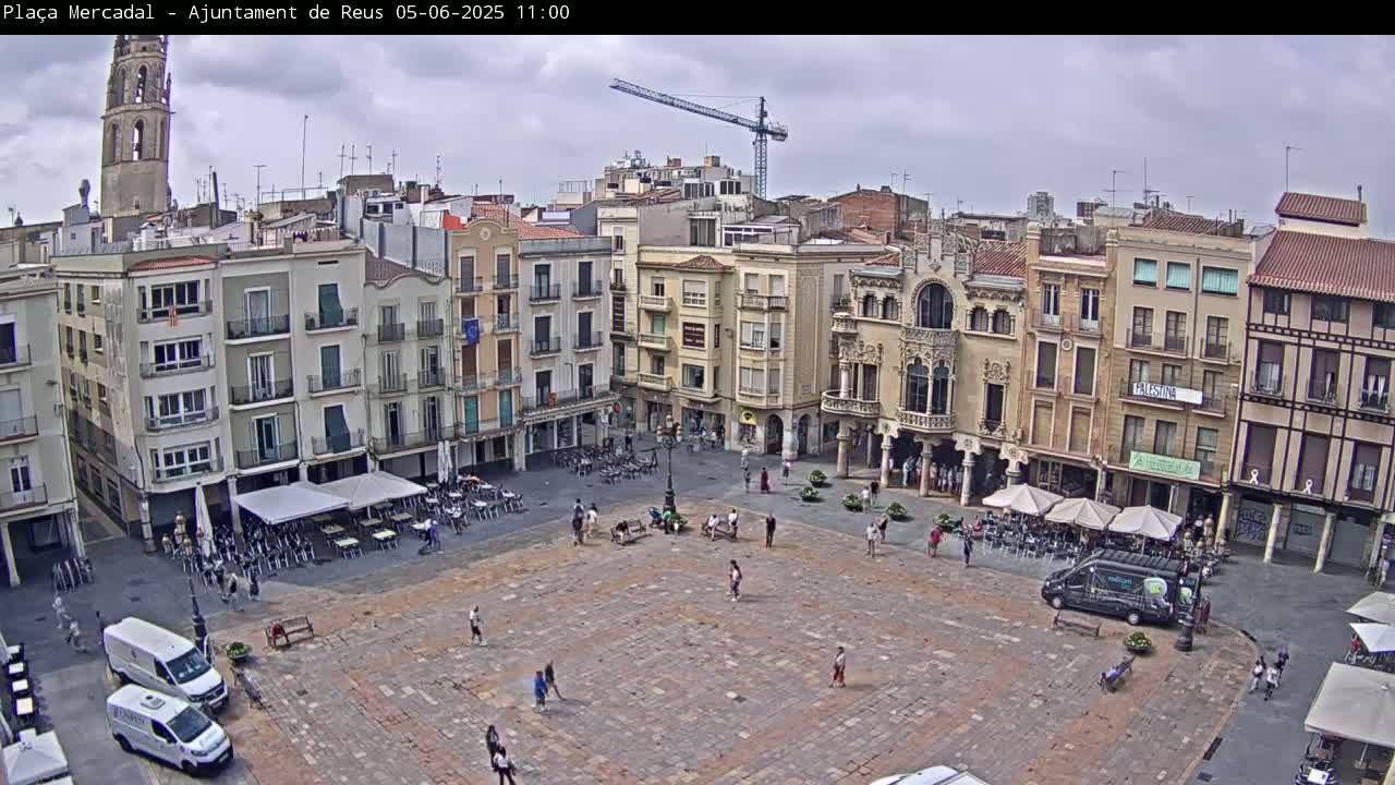 A mostly sunny, partly cloudy day overlooks a large town square with several buildings surrounding it, a few people walking around, and vehicles parked nearby.