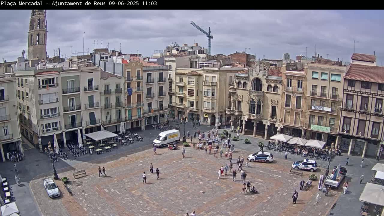 A town square with several multi-story buildings surrounding it is filled with people under a partly cloudy sky.