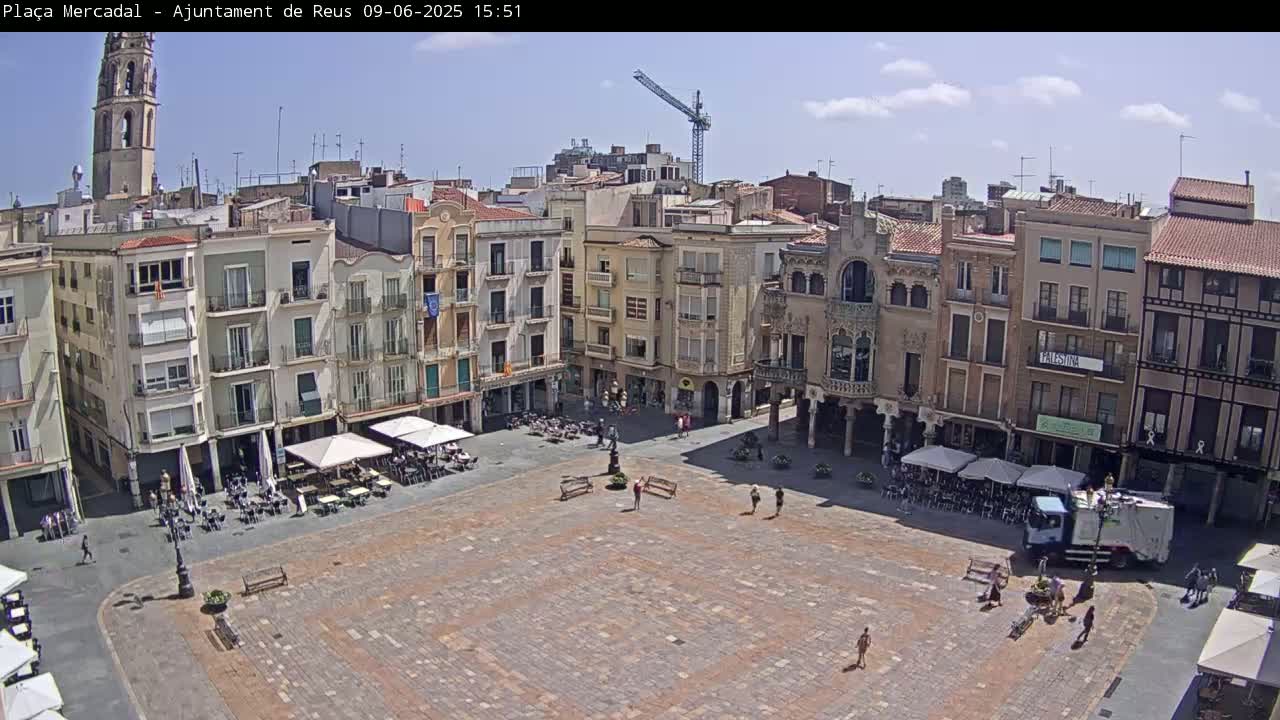 A large, paved town square with several buildings surrounding it, outdoor cafes with umbrellas, and a garbage truck, under a mostly sunny sky.