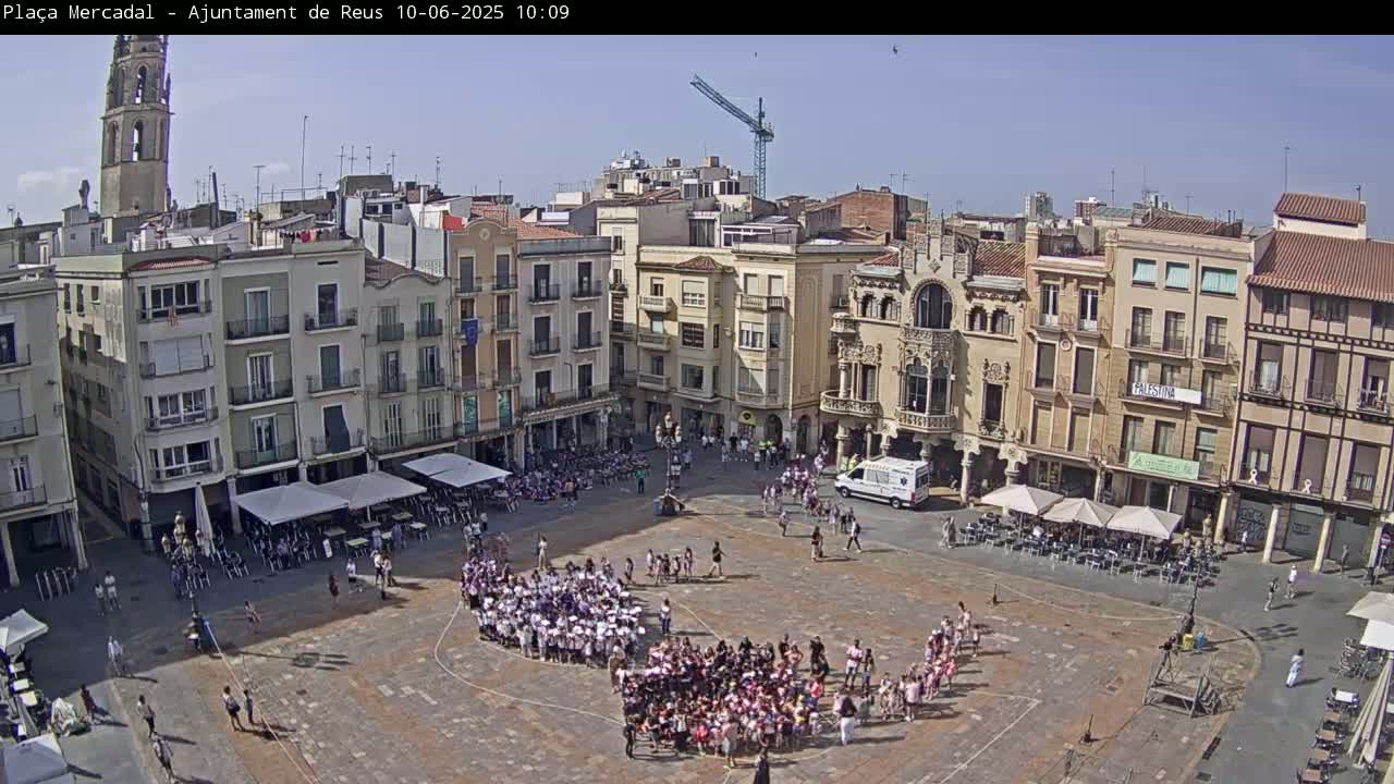 A large town square, under a clear blue sky, is filled with people, many of whom are children in groups, gathered around a central point.