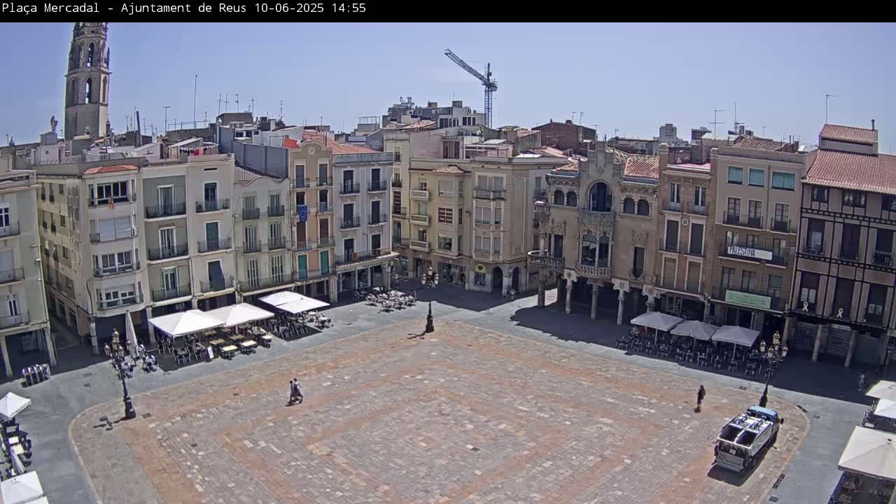A large, paved town square with outdoor seating at restaurants surrounding it, under a clear sunny sky, is viewed from above, with multi-story buildings bordering the square and a construction crane visible in the distance.