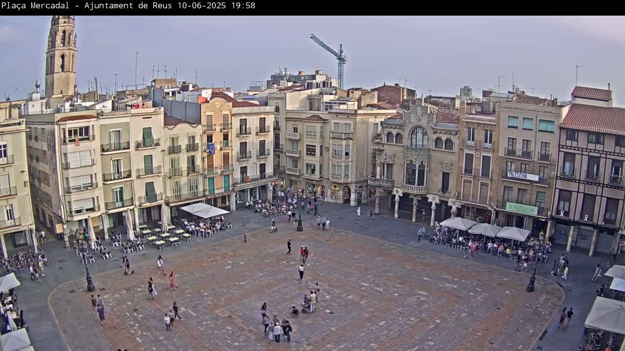 A large, mostly empty town square with surrounding buildings, outdoor seating areas, and a few pedestrians, under a mostly clear sky.