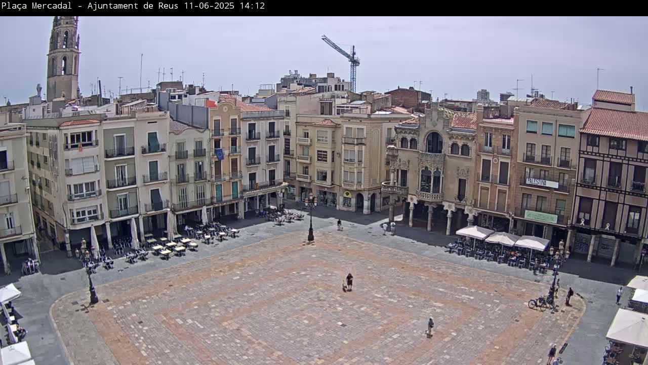 A large, mostly empty town square with surrounding buildings, outdoor cafes, and a crane visible in the distance on a partly sunny day.