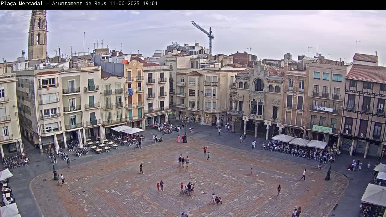 A large town square, mostly paved, is surrounded by multi-story buildings under a partly cloudy sky; people are scattered throughout the square, sitting at outdoor cafes and walking.