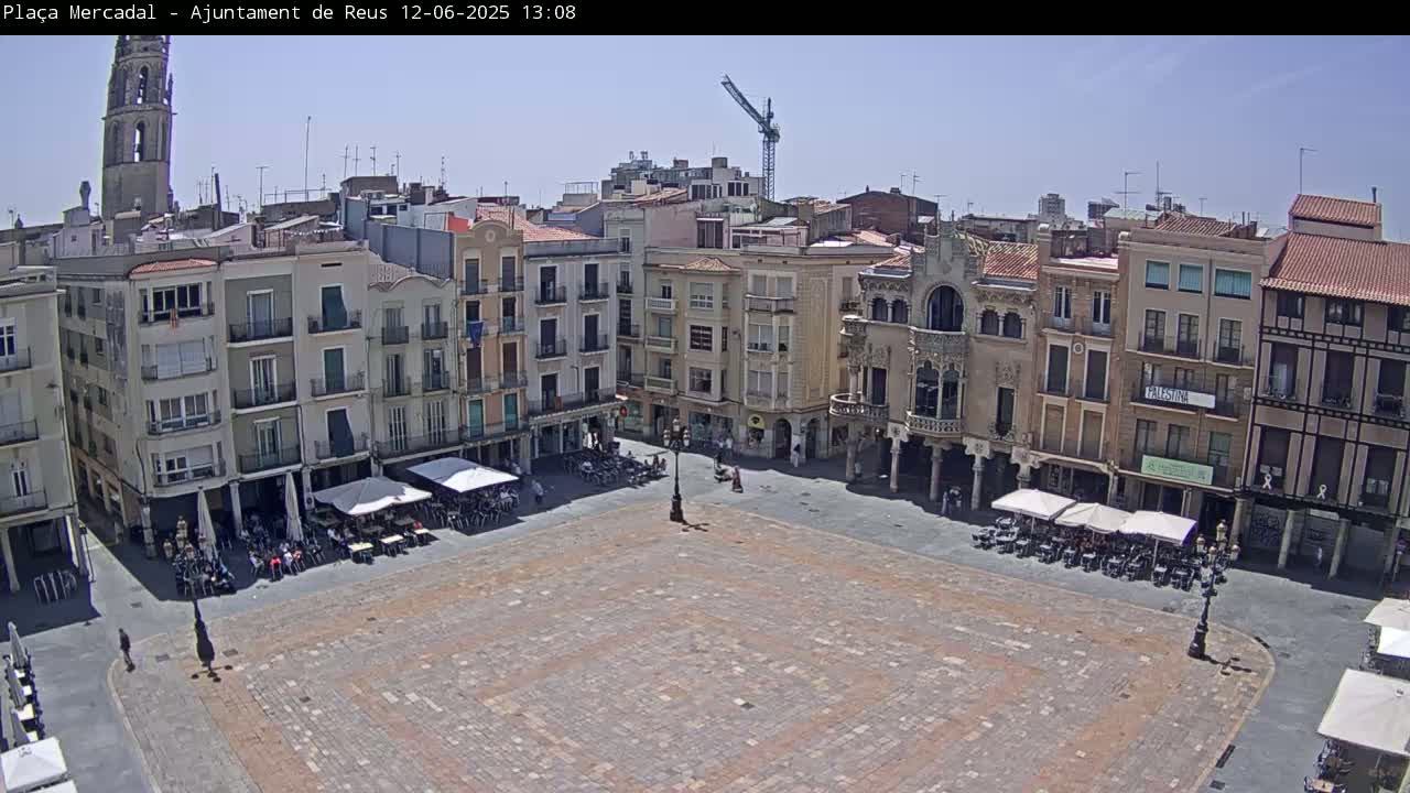 A large, paved town square is surrounded by multi-story buildings under a clear, sunny sky.