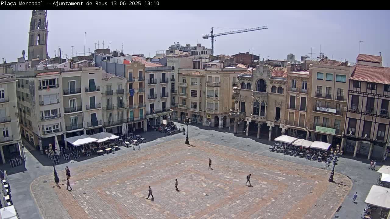 A large, mostly empty town square paved with brick is surrounded by multi-story buildings under a clear, sunny sky, with a construction crane visible in the distance.