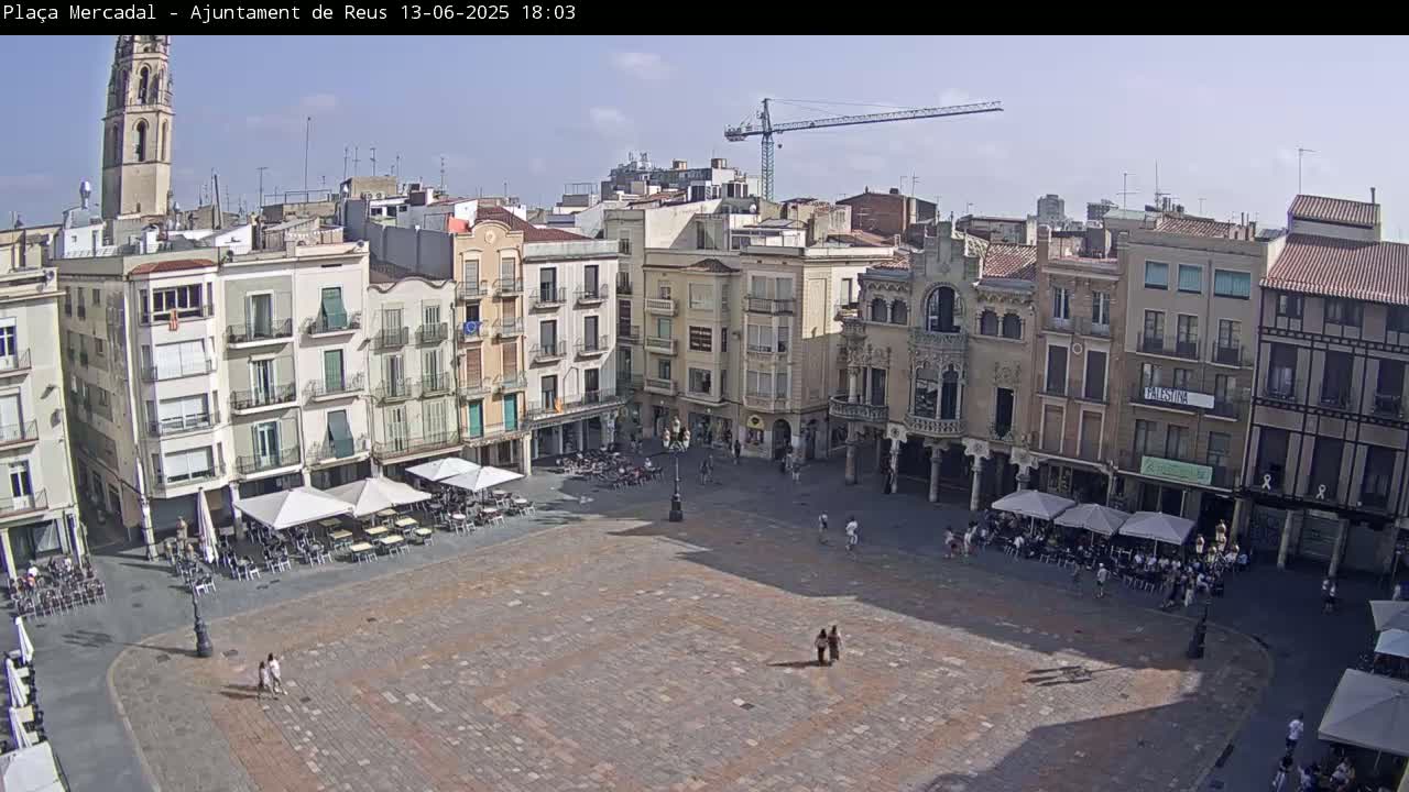 A large town square with several multi-story buildings surrounding it, outdoor cafes with patrons seated at tables, and a construction crane visible in the distance under a mostly sunny sky.