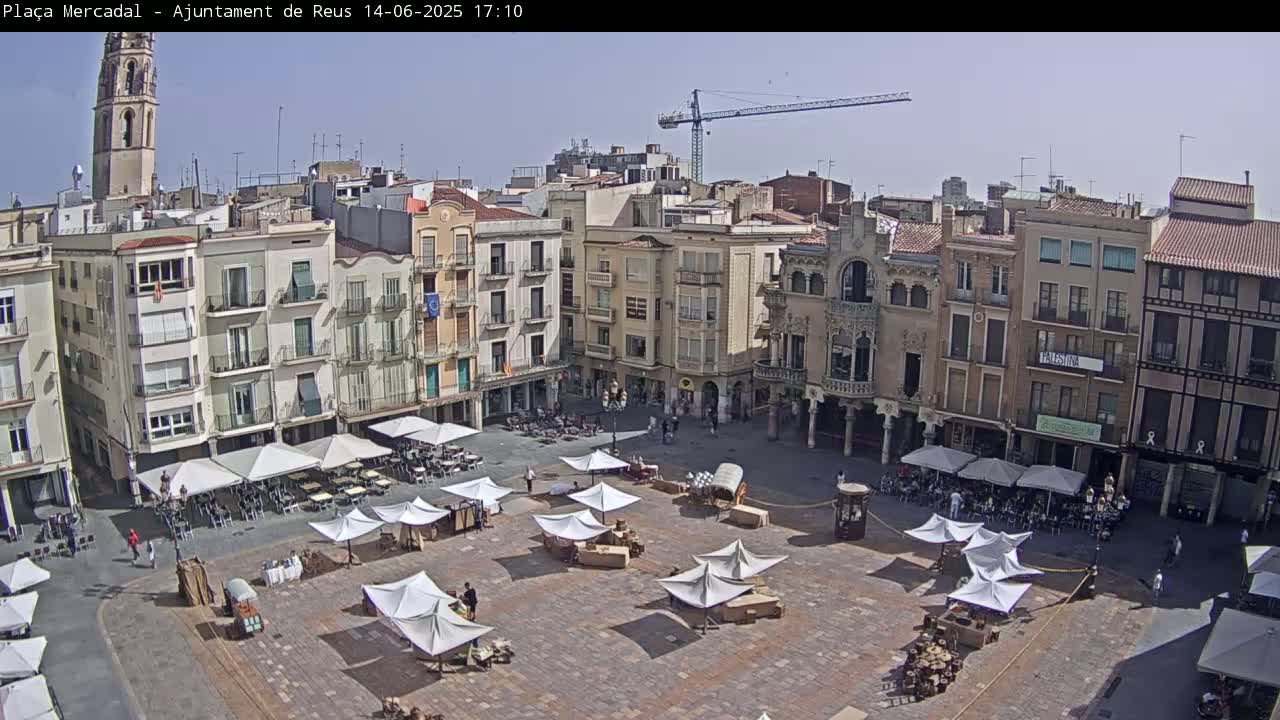 A sunny town square with several white market stalls and outdoor café umbrellas is surrounded by multi-story buildings, including a tall bell tower and a construction crane in the distance.