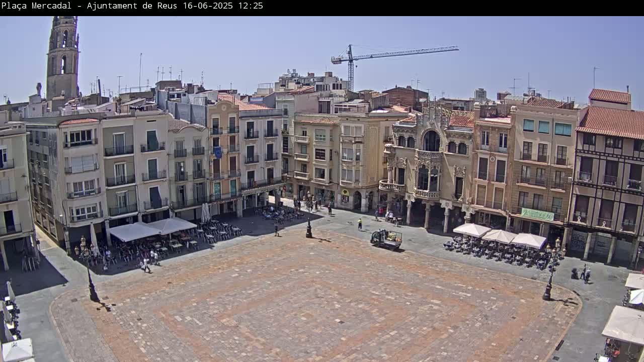A large, mostly empty town square with several buildings surrounding it, outdoor cafes with umbrellas, and a construction crane in the background on a sunny day.