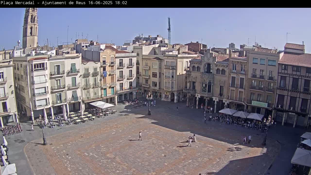 A large, mostly empty town square with several buildings surrounding it on a sunny day, some outdoor cafes are partially occupied.