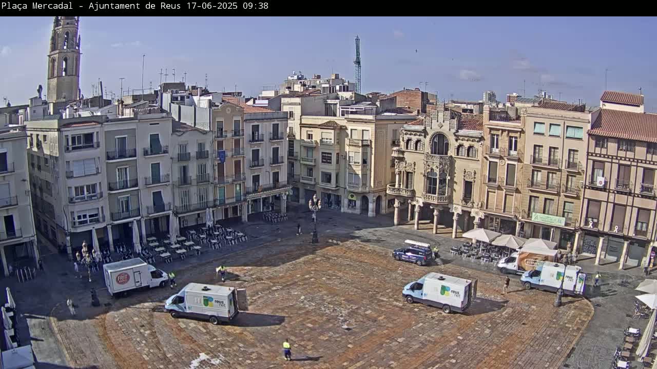 A large, mostly empty town square paved with brick is surrounded by multi-story buildings under a clear, sunny sky; several small vehicles are parked in the square.