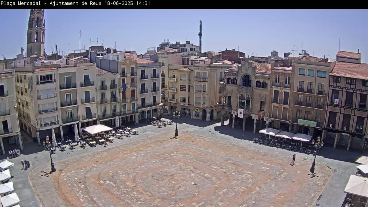 A large, mostly empty town square with surrounding buildings, outdoor cafes, and a clear sunny sky.