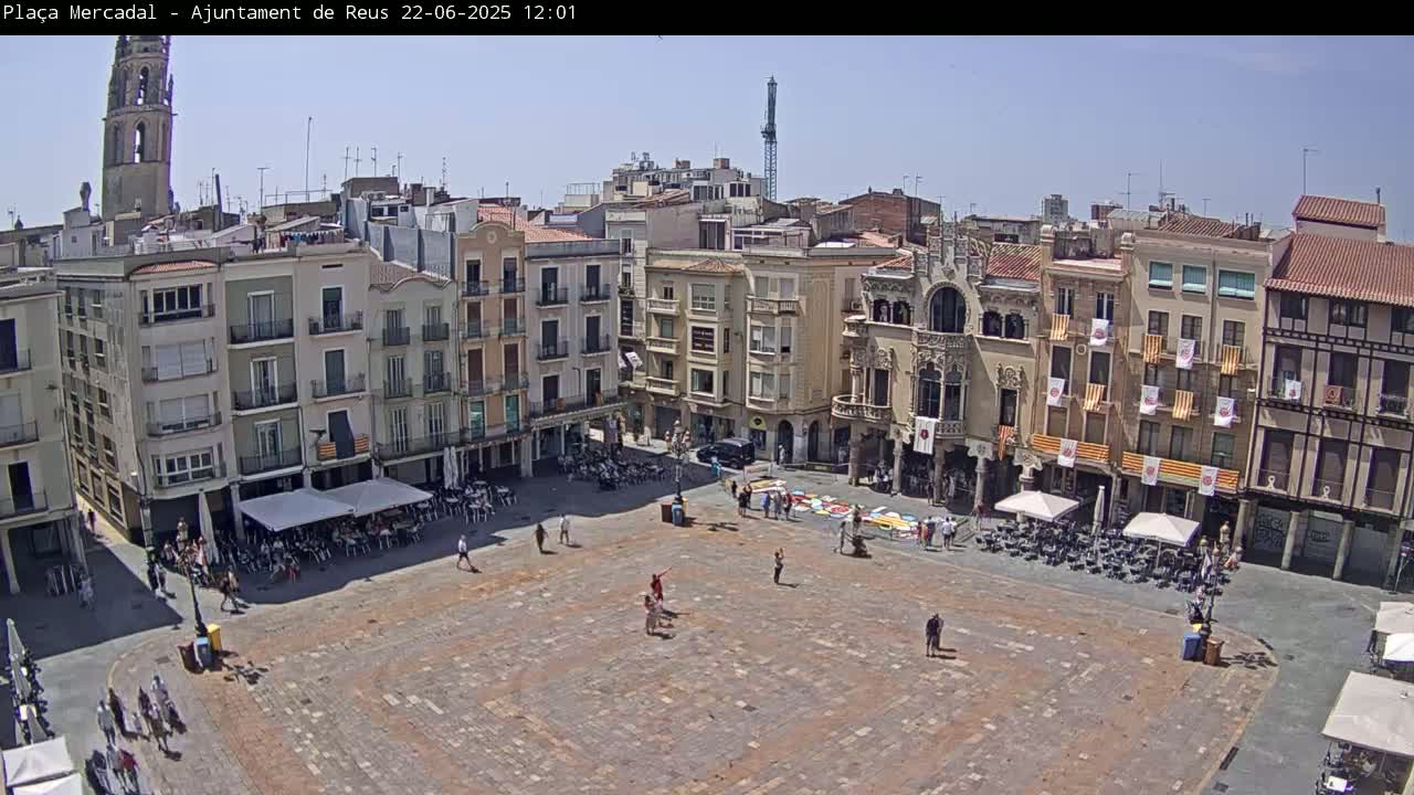 A large town square, under a clear blue sky, is surrounded by multi-story buildings, with a few people scattered throughout the square and some outdoor seating areas.