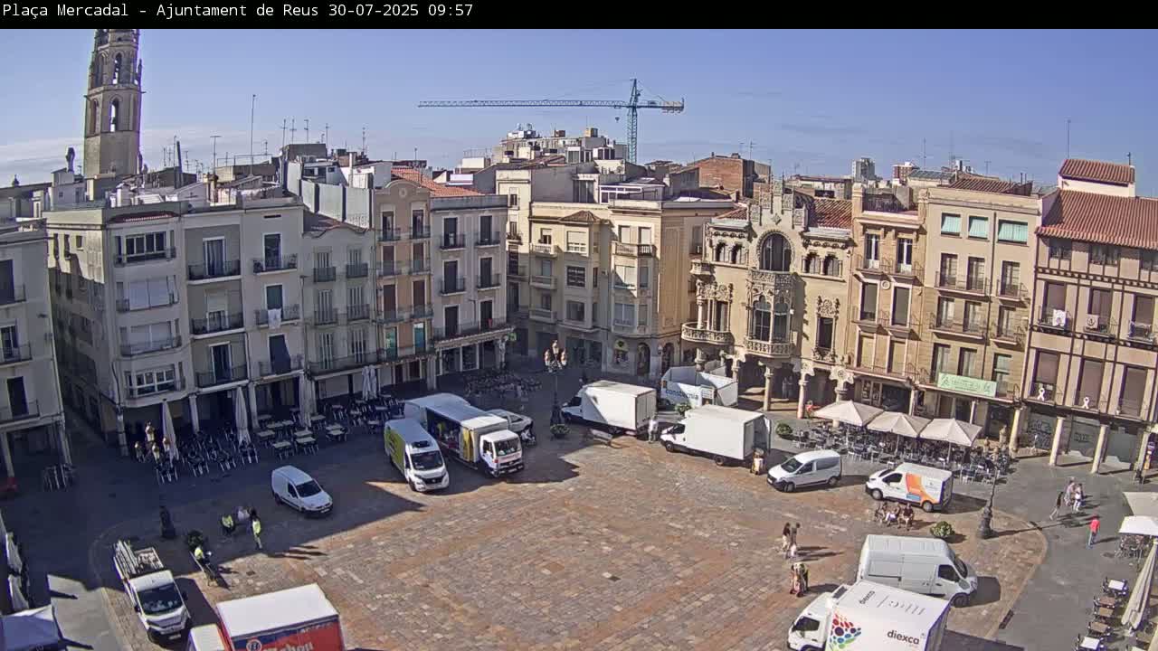 A sunny day reveals a large town square surrounded by multi-story buildings, with several delivery trucks and people scattered throughout.