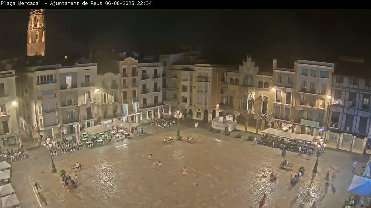 A nighttime view of a town square, wet from rain, with people sitting at outdoor cafes and children playing in the square, all surrounded by illuminated buildings.