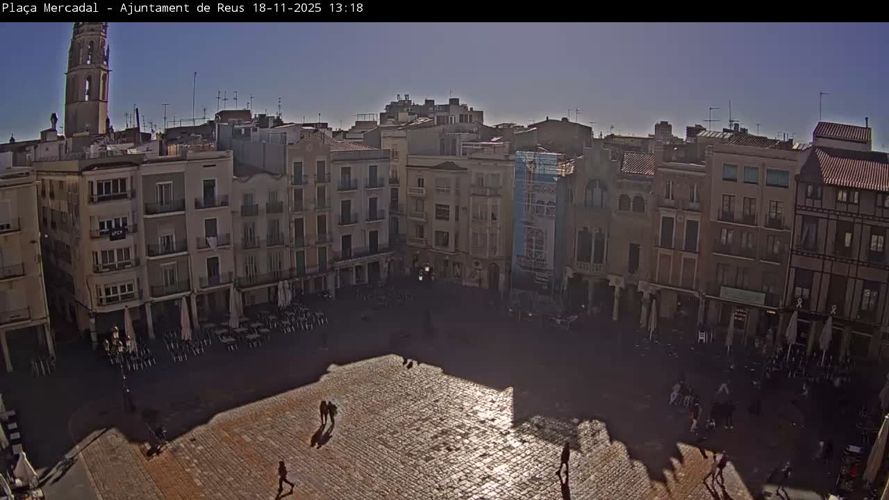 The image shows a wet, illuminated city plaza at night surrounded by multi-story buildings with active outdoor cafes and a few pedestrians, indicating recent rain.