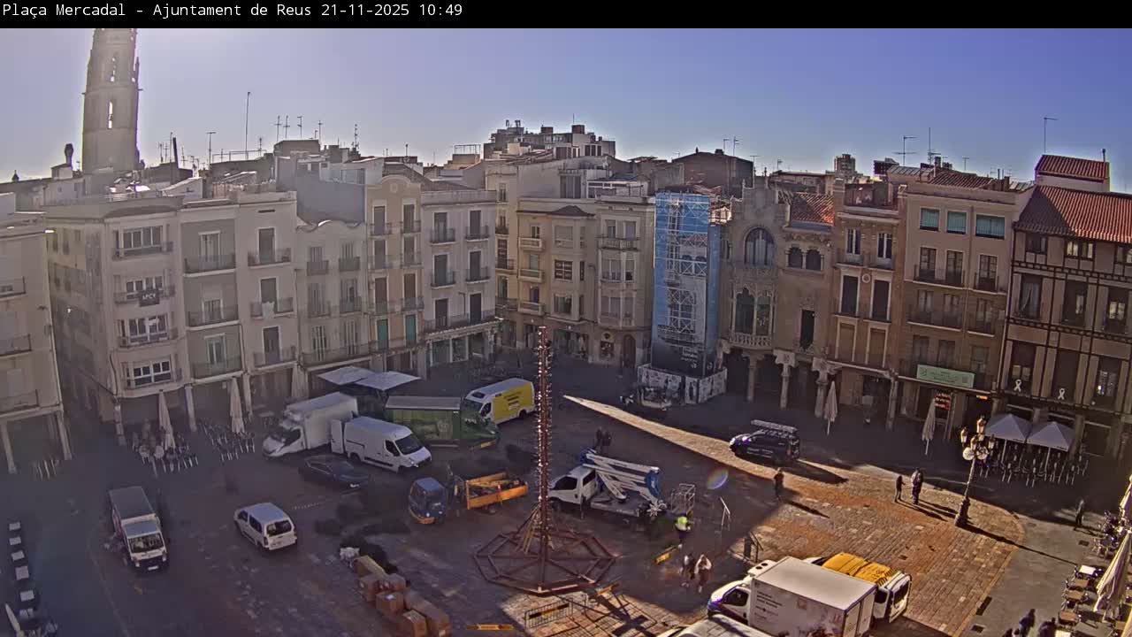 Under a bright and clear morning sky, an active town square with historic multi-story buildings features vehicles, pedestrians, and a large decorative structure being assembled in the center, with a tall church tower visible in the background.