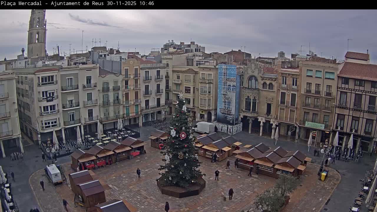 An overcast day overlooks a festive town square featuring a tall decorated Christmas tree surrounded by market stalls, numerous multi-story buildings, and a distant church tower, with several pedestrians walking throughout the scene.