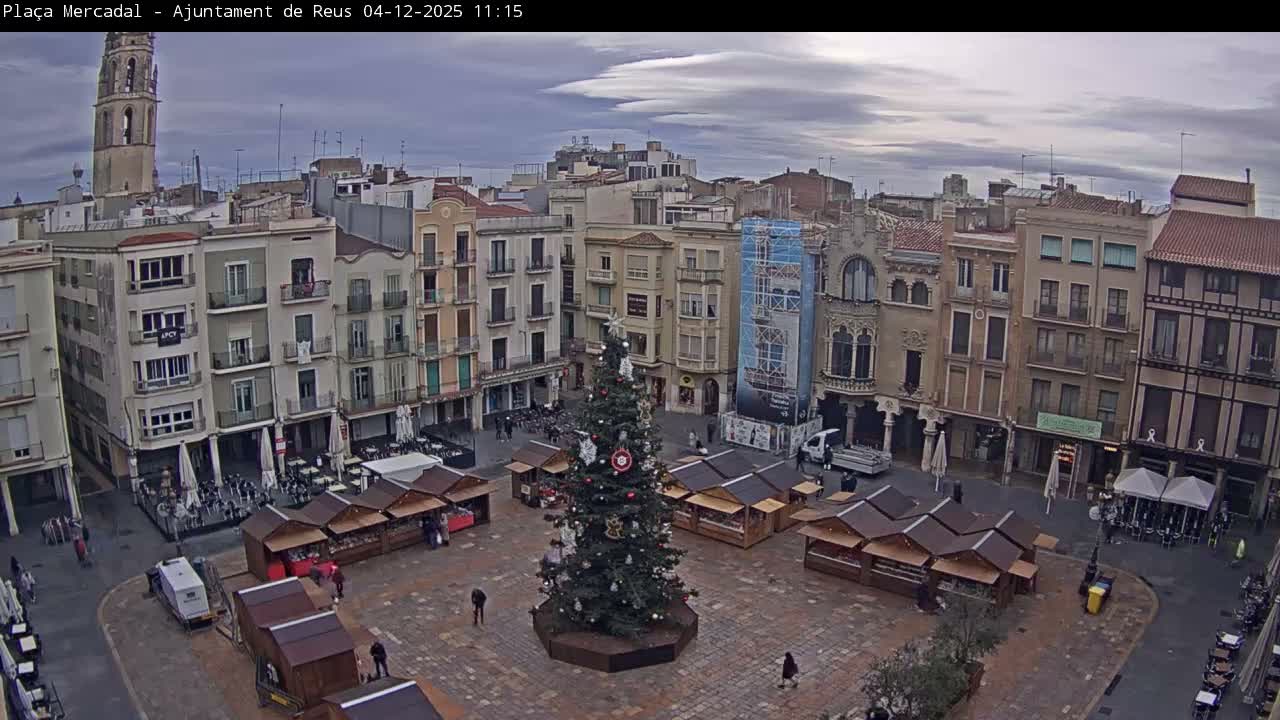 An outdoor plaza is decorated for a Christmas market with a large tree in the center surrounded by numerous stalls, all framed by multi-story buildings and a prominent church tower under a cloudy, overcast sky.