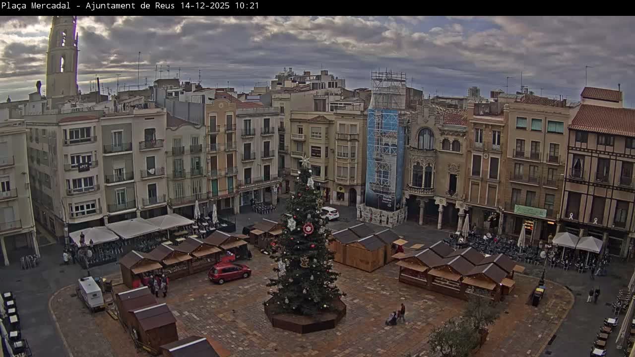 An outdoor plaza is decorated for a Christmas market with a large tree in the center surrounded by numerous stalls, all framed by multi-story buildings and a prominent church tower under a cloudy, overcast sky.