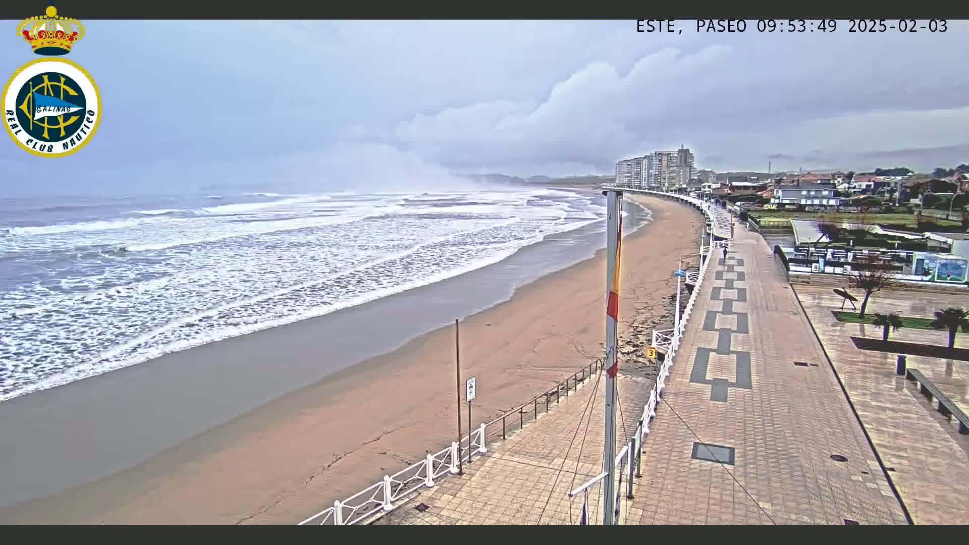 A sandy beach with moderate waves under a cloudy sky is bordered by a paved walkway and buildings.