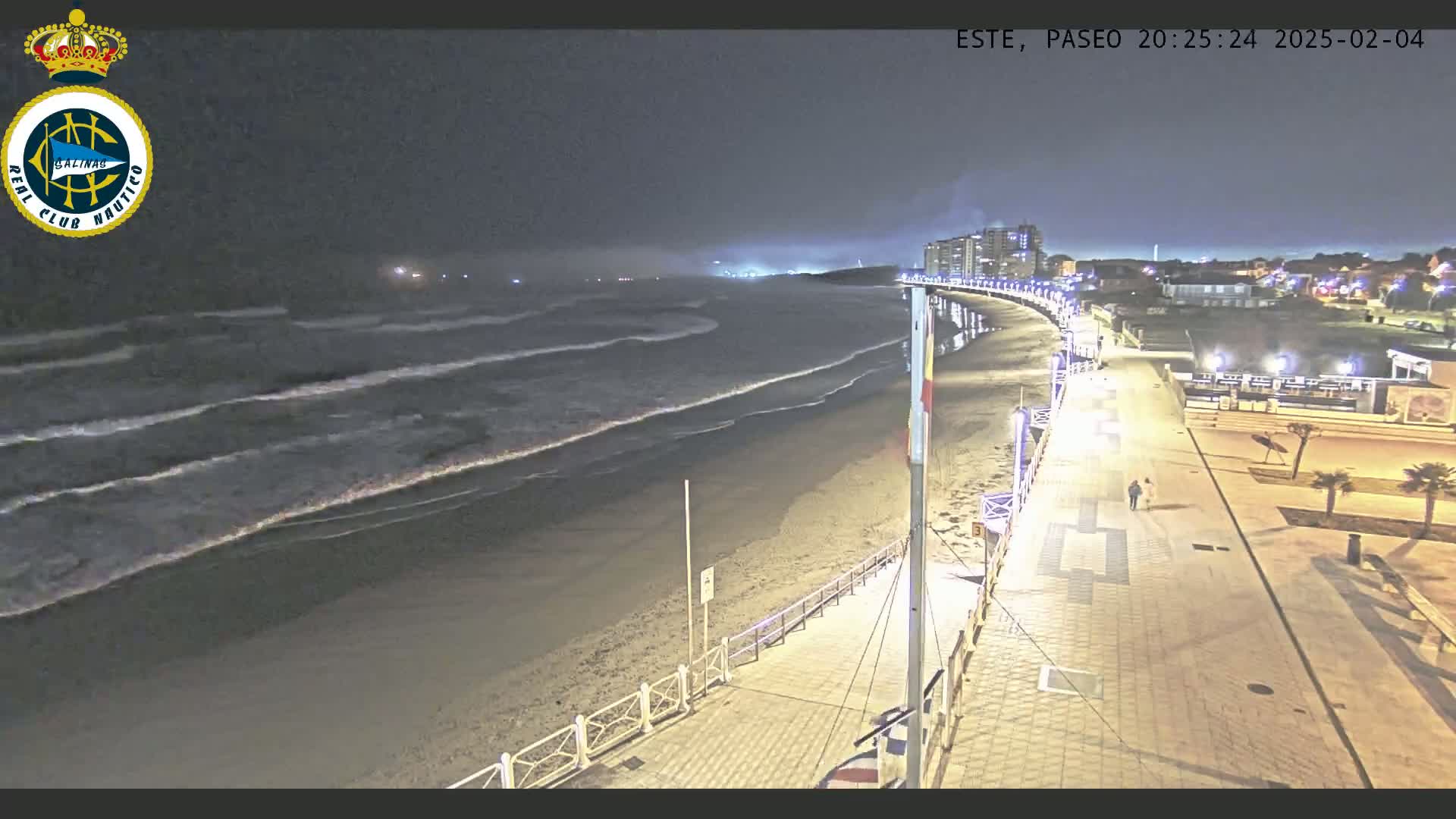 A nighttime view shows a dark, sandy beach with moderate waves under a mostly clear sky, alongside a brightly lit boardwalk with a couple walking on it.