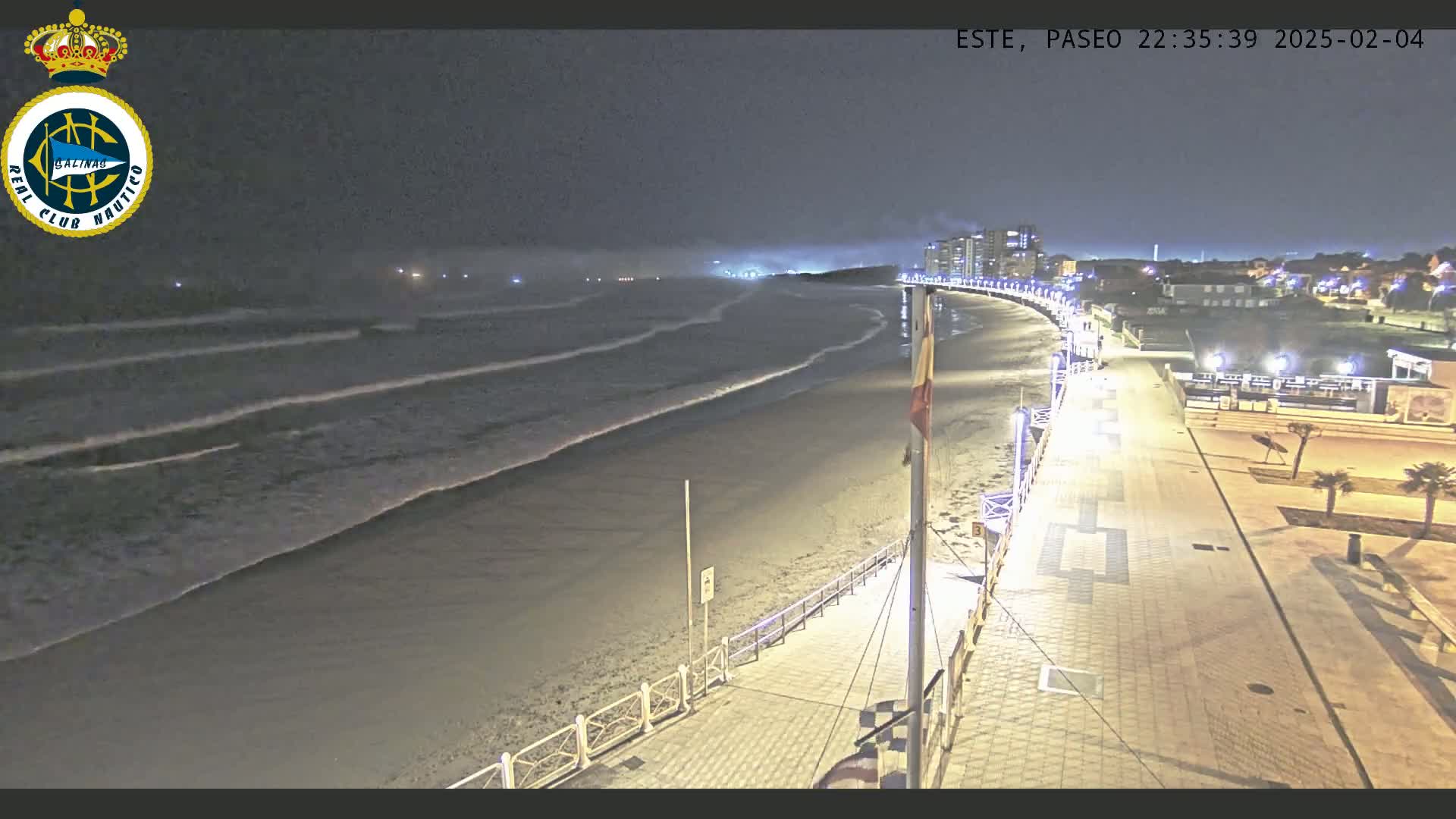 A nighttime, high-angle view shows a mostly empty beach with gentle waves lapping the shore, next to a paved walkway lined with lights, leading to buildings in the distance under a cloudy sky.