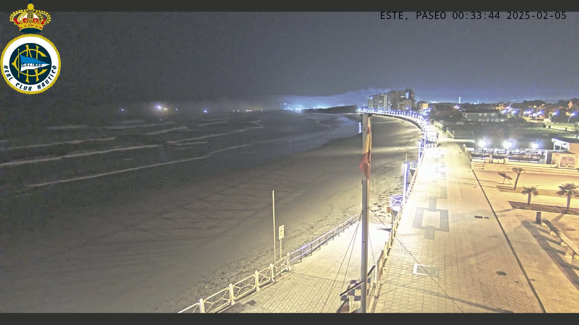 A nighttime view of a beach with gentle waves, a paved walkway lined with lights, and buildings in the distance under a dark sky.