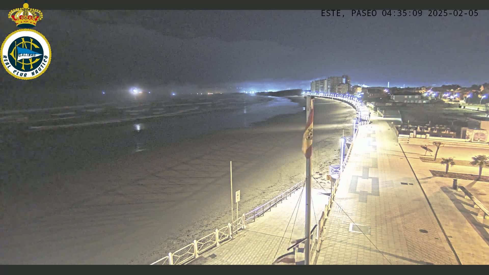 A nighttime view of a mostly empty beach and boardwalk, with calm waves and lights illuminating the path.