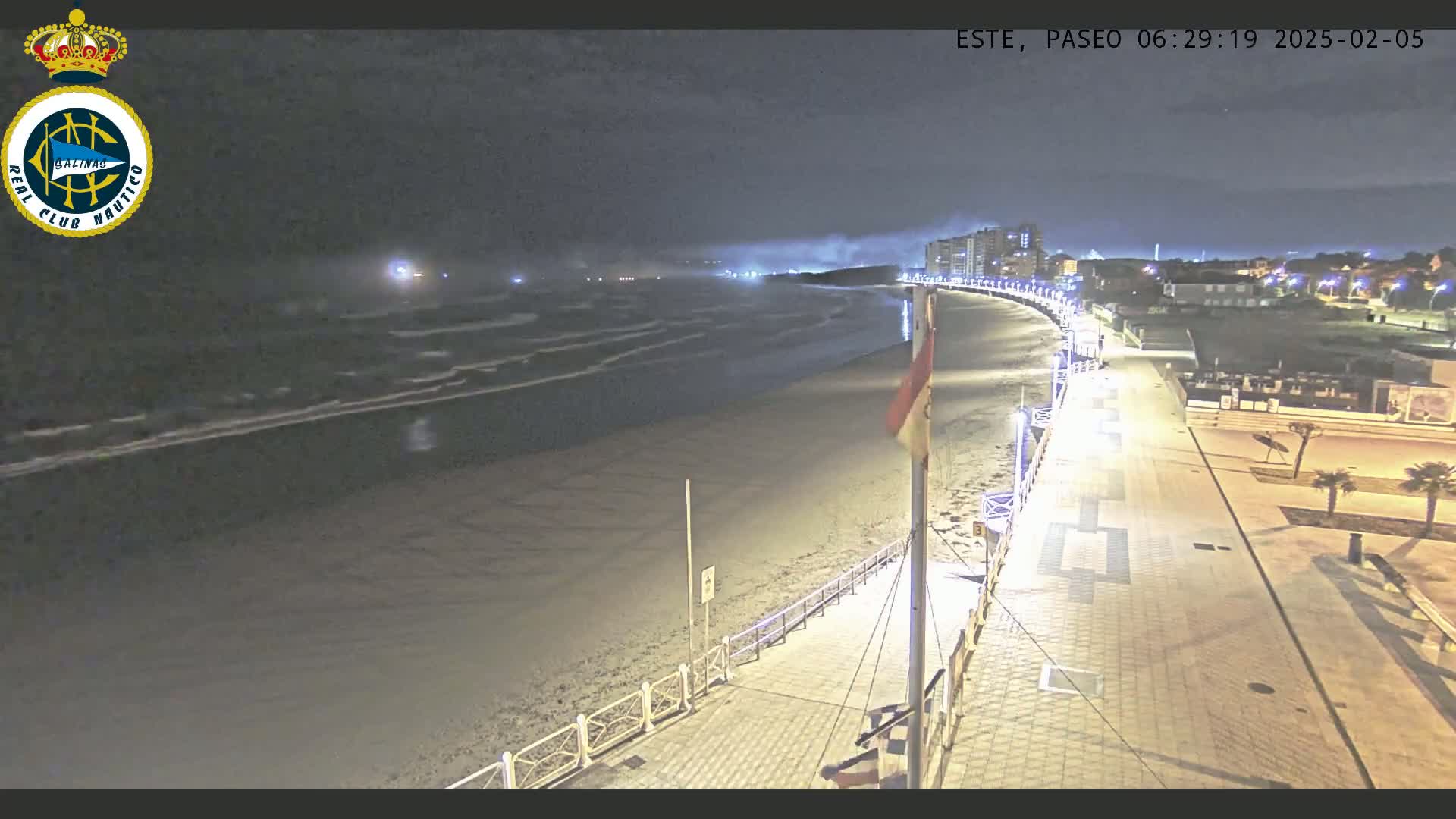 A nighttime beach scene shows a paved walkway lit by streetlights next to a sandy beach with gently lapping waves under a mostly cloudy sky.